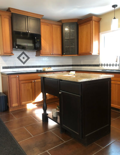 A kitchen with wooden cabinets, a dark-colored island, granite countertops, a built-in microwave, tile backsplash with diamond accents, and large brown floor tiles. Sunlight shines through a nearby window.